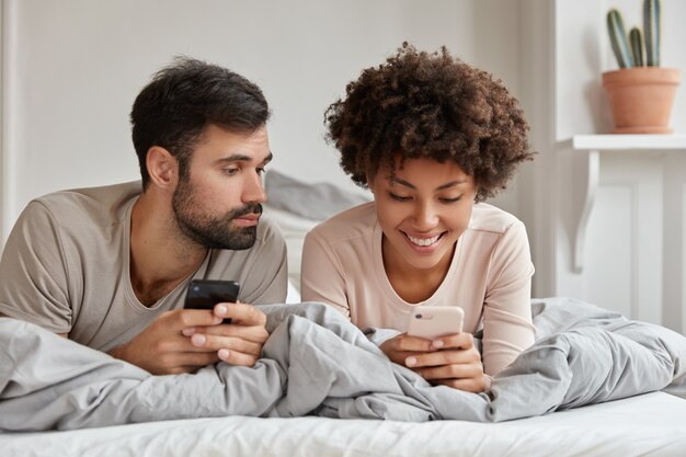 Lovers enjoy leisure time at home, hold modern smart phones in hands, read income notification, lie in bed, rejoice good news, chat in network, connected to high speed internet, browse web page