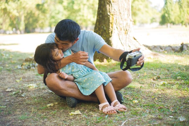 Loving Asian father kissing his child for camera. Happy man sitting on grassy ground with daughter on his knees hugging kissing her cheek, taking their photos. Fatherhood, modern technologies concept