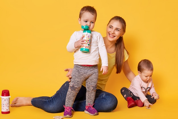 loving attractive mother looks after her little children, twins playing with mommy. Playful kids drink tasty baverage from her bootle while her sister eating coockies. Infants are hungry.