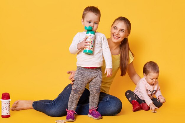 loving attractive mother looks after her little children, twins playing with mommy. Playful kids drink tasty baverage from her bootle while her sister eating coockies. Infants are hungry.