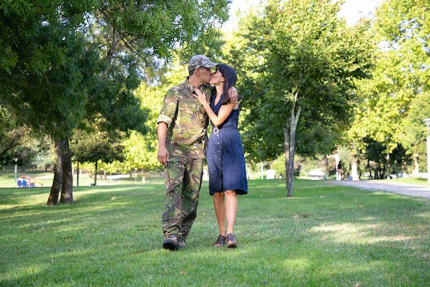 Loving Caucasian couple embracing, kissing and walking together on lawn in park. Middle-aged soldier in military uniform, hugging his pretty wife. Family reunion, weekend and returning home concept