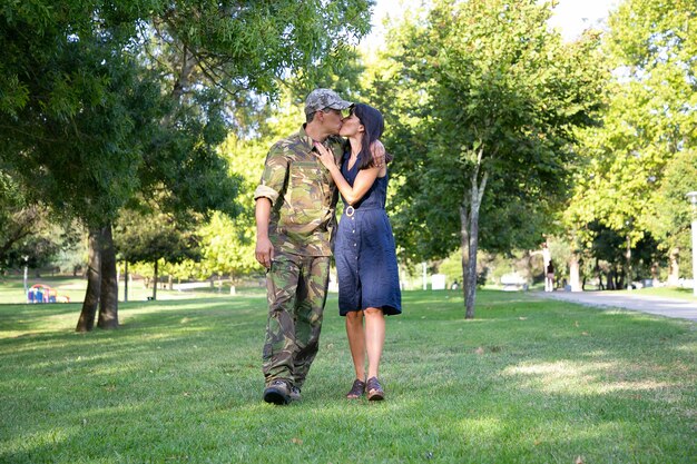 Loving Caucasian couple embracing, kissing and walking together on lawn in park. Middle-aged soldier in military uniform, hugging his pretty wife. Family reunion, weekend and returning home concept