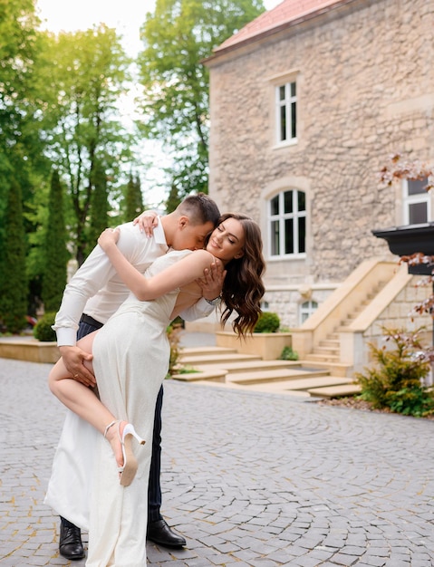 Loving couple of bridal man hugging and kissing bride in elegant long white wedding dress Attractive groom and bride brunette girl posing outdoors near ancient building Wedding happy day Engagement