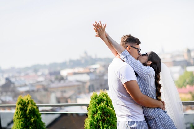 Loving couple bride with groom in pajamas kissing while standing on balcony on city background Brides in sunglasses hugging enjoying happy moment Woman with veil in hairstyle