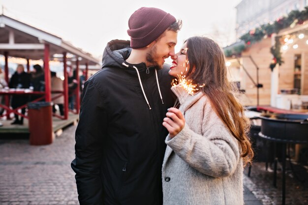 Loving couple embracing in amusement park in winter weekend. Happy black-haired girl celebrating christmas with boyfriend and posing in front of carousel in cold day..