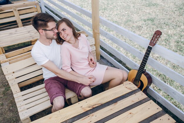 Любить пара охватывающей на деревянной скамейке с гитарой рядом с ним