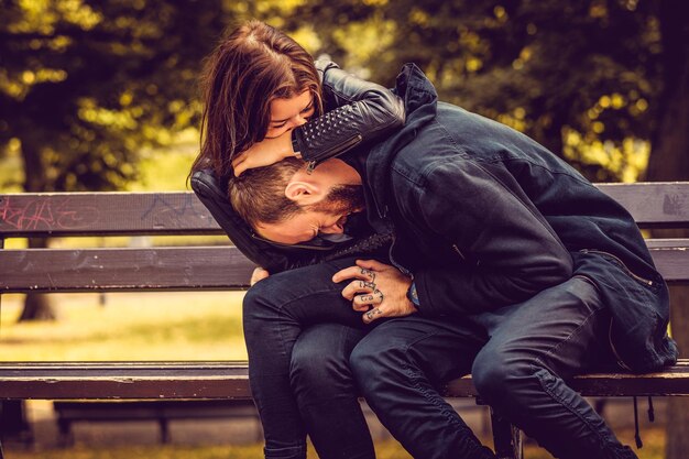 Loving couple posing on a bench in autumn city park. Bearded male hugging attractive brunette female.