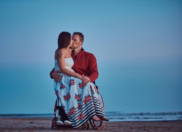 Влюбленная пара, женщина сидит на коленях у мужа и целуется на пляже на фоне яркого рассвета.