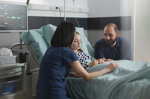 Loving family comforting under treatment sleeping ill kid in hospital pediatric ward. Considerate parents sitting beside hospitalized sick little daughter inside children healthcare facility room.