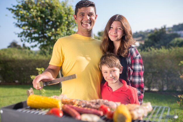 子供と一緒に肉や野菜を焼く愛情のある父。幼い息子と10代の娘と一緒にバーベキューグリッドの近くに立っている黄色のTシャツを着た黒髪の男。バーベキュー、料理、食べ物、家族のコンセプト