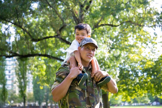 首に息子を抱き、都市公園を歩いている愛情のある父。制服を着たお父さんの首に座って、彼を抱き締めて、目をそらしている幸せな白人の息子。家族の再会、父性、帰国の概念