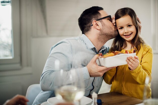 彼女がダイニングテーブルで昼食時に彼に食べ物を提供している間、彼の小さな娘にキスする愛情のある父親