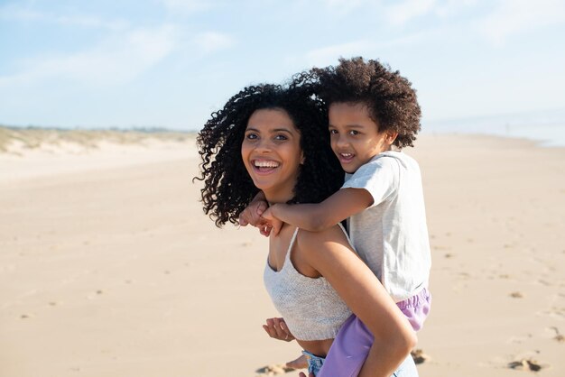 사랑하는 엄마와 딸이 해변에서 시간을 보내고 있습니다. 아프리카계 미국인 가족은 걷고, 웃고, 놀고, 등에 타고 있습니다. 여가, 가족 시간, 부모 개념