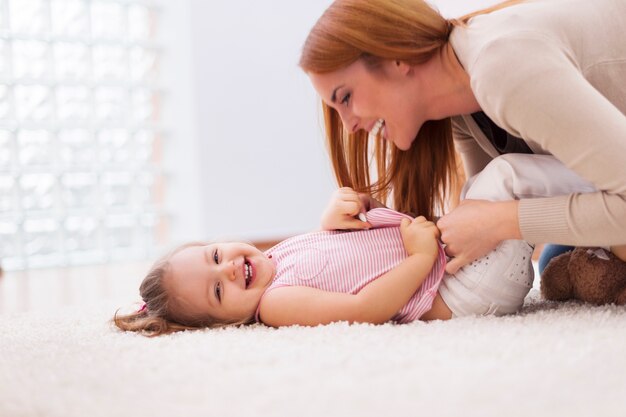 Любящая мать щекочет свою маленькую девочку на ковре дома