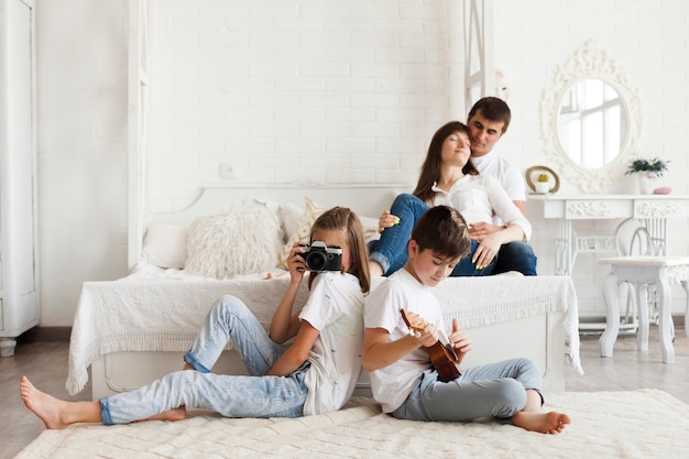 Loving parent sitting on bed and their daughter holding camera and son playing ukulele