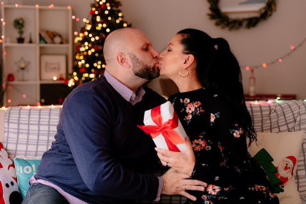 любящие романтические муж и жена дома на Рождество, сидя на диване в гостиной, жена держит подарочный пакет, целуется в губы