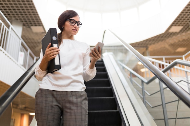 Low angle of businesswoman with smartphone and binder on escalator