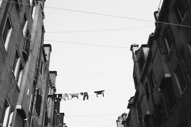 Low angle greyscale shot of clothes hanging on the cable between high concrete buildings