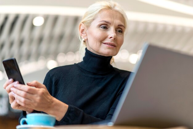 Low angle of older business woman working on laptop with smartphone