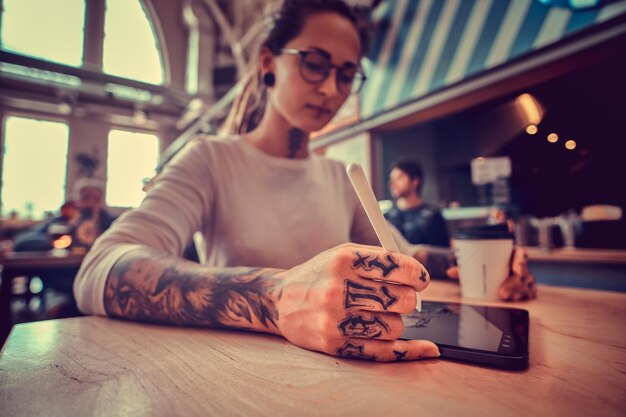 Low angle photo of attractive pensive girl with dreadlocks and tattooes. Dhe is sketching while sitting at cafe.