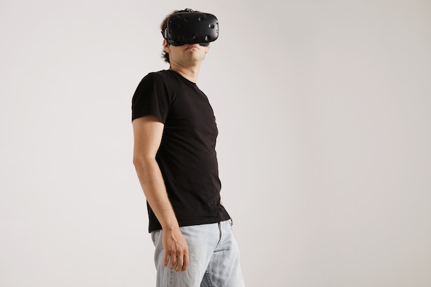 Low angle portrait of a man wearing VR headset, blank black t-whirt and jeans looking around isolated on white