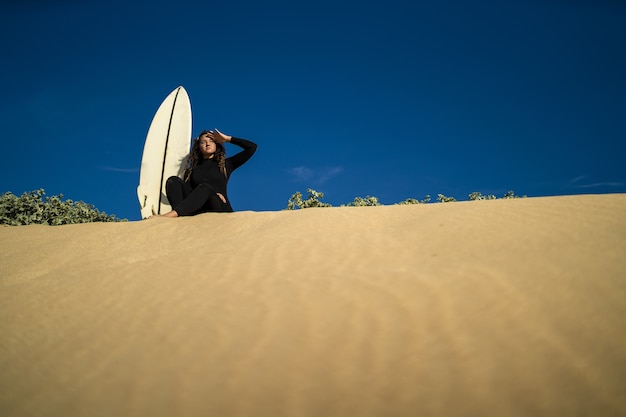 側面にサーフボードを置いて砂丘に座っている魅力的な女性のローアングルショット