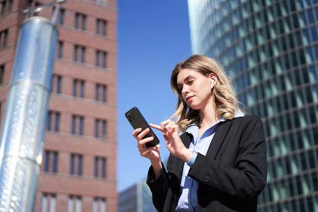 Low angle shot of businesswoman standing on street on sunny day holding smartphone and using app tex
