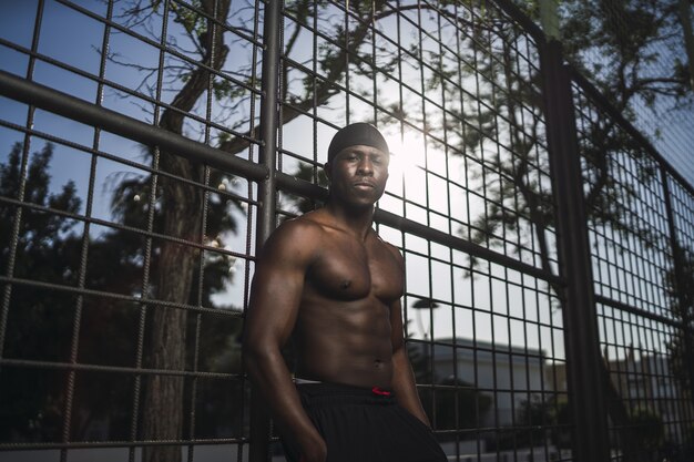 Low angle shot of a half-naked African-American male leaning on the fence at the basketball court