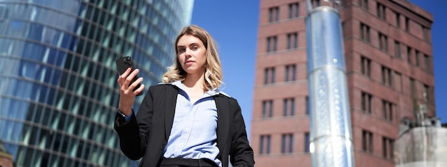 Бесплатное фото Низкоугольный снимок деловой женщины в костюме, стоящей на улице и смотрящей на мобильный телефон
