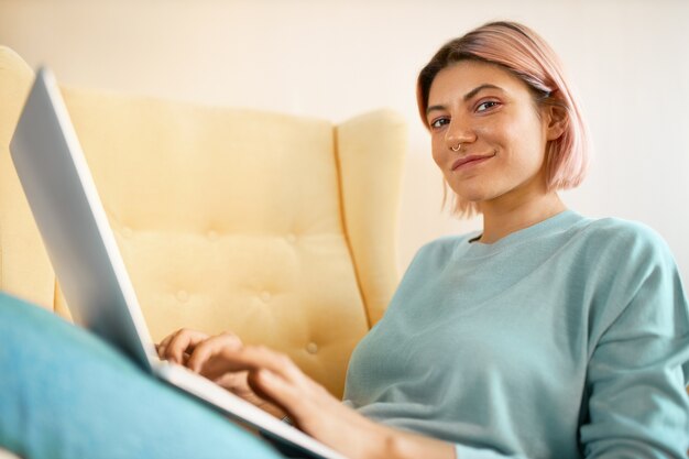 Low angle view of charming stylish student girl doing homework using generic laptop, sitting on sofa, keyboarding, using high speed wireless internet connection. Technology and electronic gadgets