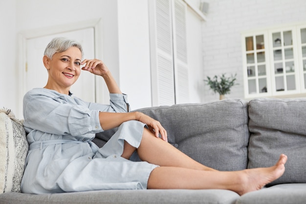 Low angle view of fashionable elegant mature sixty year old Caucasian woman with short pixie hairstyle relaxing at home sitting on gray sofa in her spacious cozy clean living room, smiling