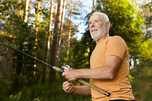 楽しい表情をした濃い灰色のひげを持つハンサムな高齢者60歳の男性の低角度のビュー、湖で釣りをしながら水から魚を引き出し、夏の朝を屋外で過ごす