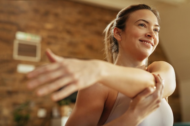 Low angle view of happy athletic woman warming up and stretching her arm at home.