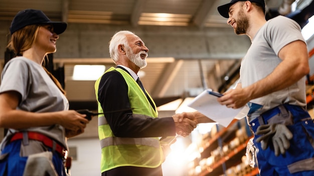 Low angle view of happy senior manager greeting his employees in a factory warehouse Me are shaking hands