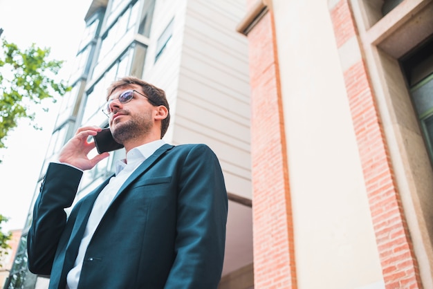 Бесплатное фото Взгляд низкого угла бизнесмена стоя под зданием говоря на мобильном телефоне