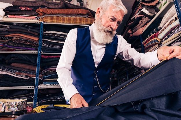Low angle view of senior male tailor taking measurement of blue fabric with measuring tape