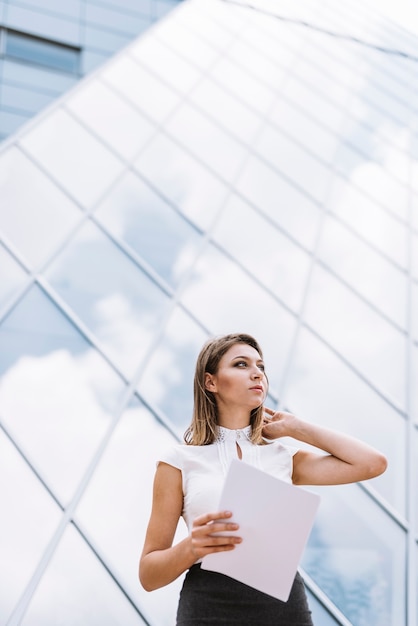 Free photo low angle view of young businesswoman standing near corporate building holding documents