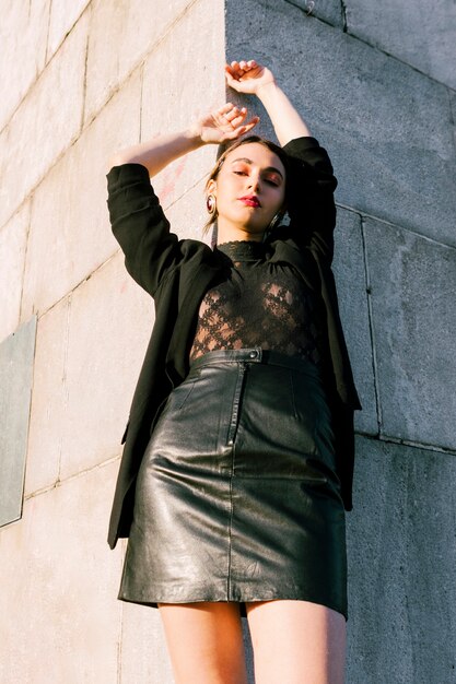 Low angle view of young fashionable woman leaning on the wall with her hand raised