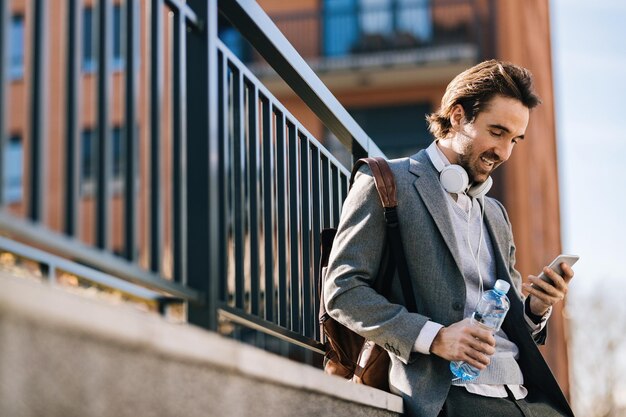 Low angle view of young happy businessman typing text message on mobile phone in the city.