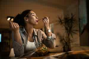 Free photo low angle view of young woman eating salad with her eyes closed while using computer at night at home