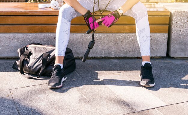 Low section of fitness young woman sitting on bench holding skipping rope in hand