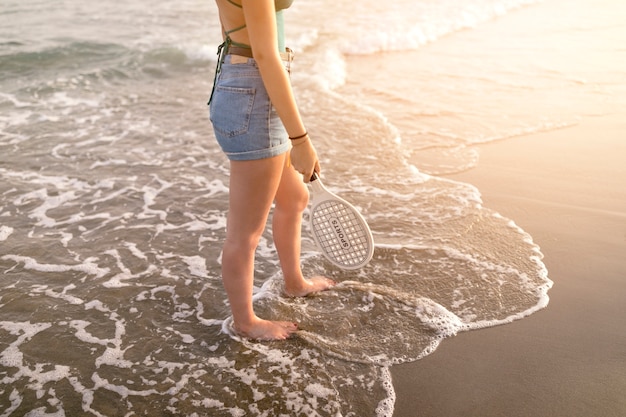 Sezione bassa della donna che tiene in mano la racchetta in piedi a piedi nudi sul litorale