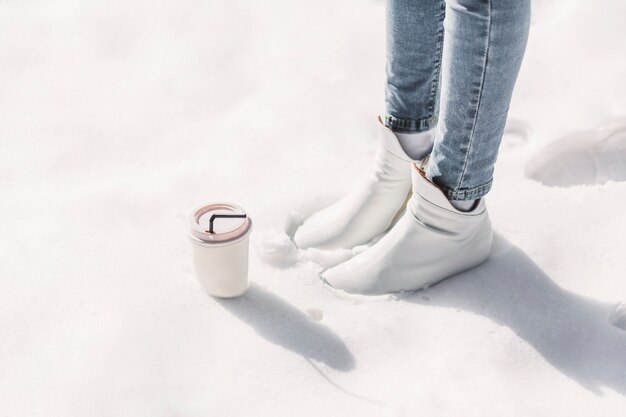 雪の上に立っているテイクアウトコーヒーカップを持つ女性の低いセクション