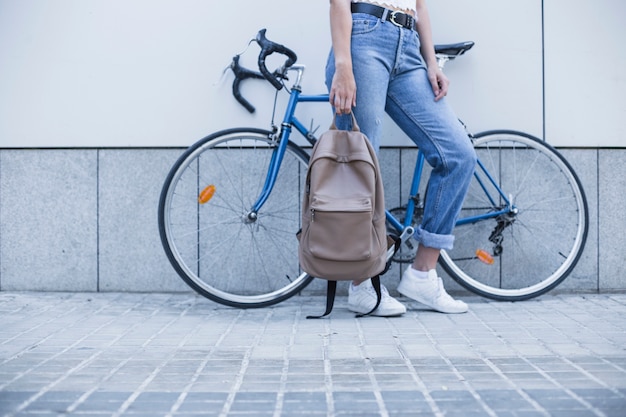 Низкий раздел молодой женщины, стоящей рядом с велосипедом, держит рюкзак
