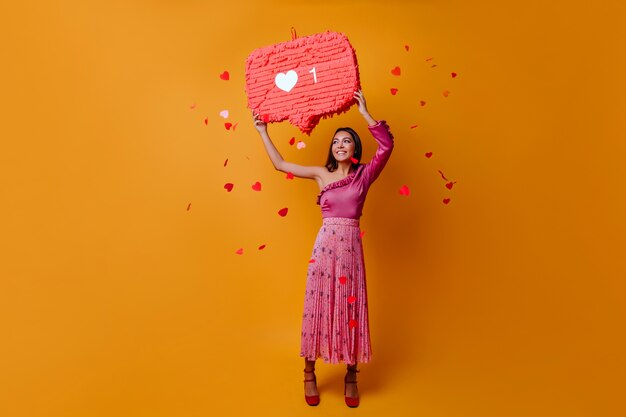 Lucky, charming woman of 23 years holds sign in form of like from Instagram and poses in full growth on orange wall with confetti