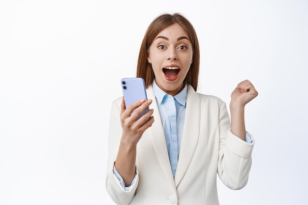 Счастливая корпоративная женщина кричит от радости, выигрывает на мобильном телефоне, торжествует от хороших новостей в Интернете, держит мобильный телефон и прыгает от счастья, белая стена