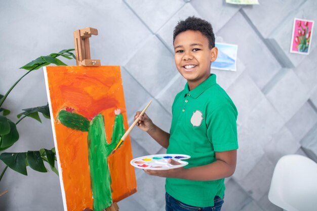 Удачный день. Темнокожий симпатичный мальчик школьного возраста в зеленой футболке с палитрой и кистью в руках возле мольберта стоит, улыбаясь в камеру в комнате