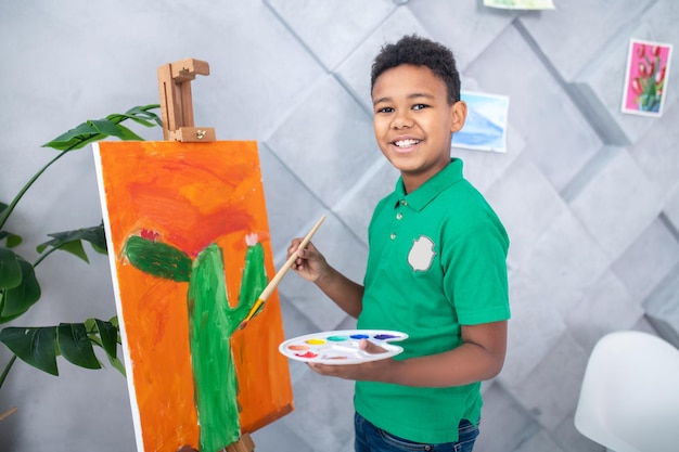 Lucky day. Dark-skinned cute school-age boy in green tshirt with palette and brush in his hands near easel standing smiling at camera in room