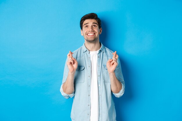 Lucky guy praying and making wish with crossed fingers, looking up with pleading face, standing against blue background