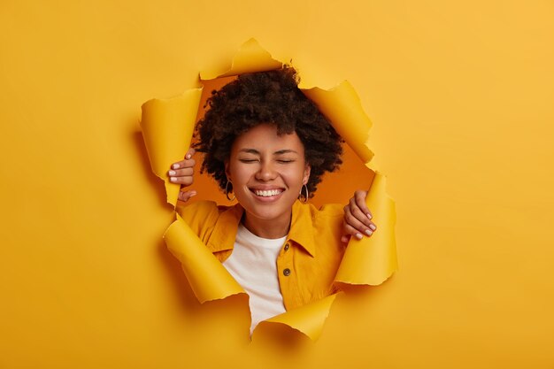 Счастливая счастливая афроамериканка широко улыбается, у нее беззаботное настроение, одетая в модную одежду, позирует на желтом бумажном фоне
